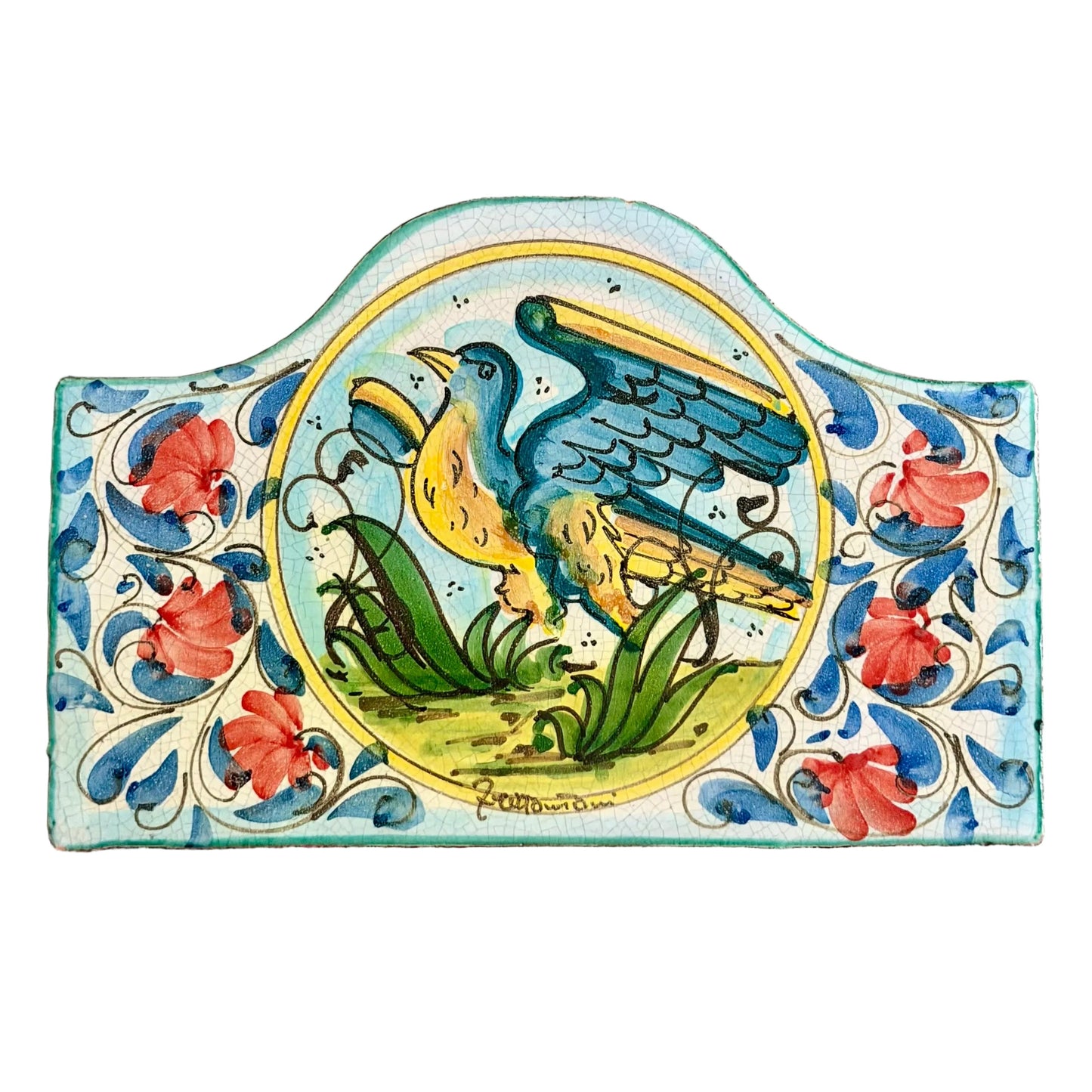 A hand-painted ceramic tile featuring a blue bird with yellow wings perched on green grass, surrounded by red flowers and blue scrollwork on a light blue background, displayed on a red patterned rug.